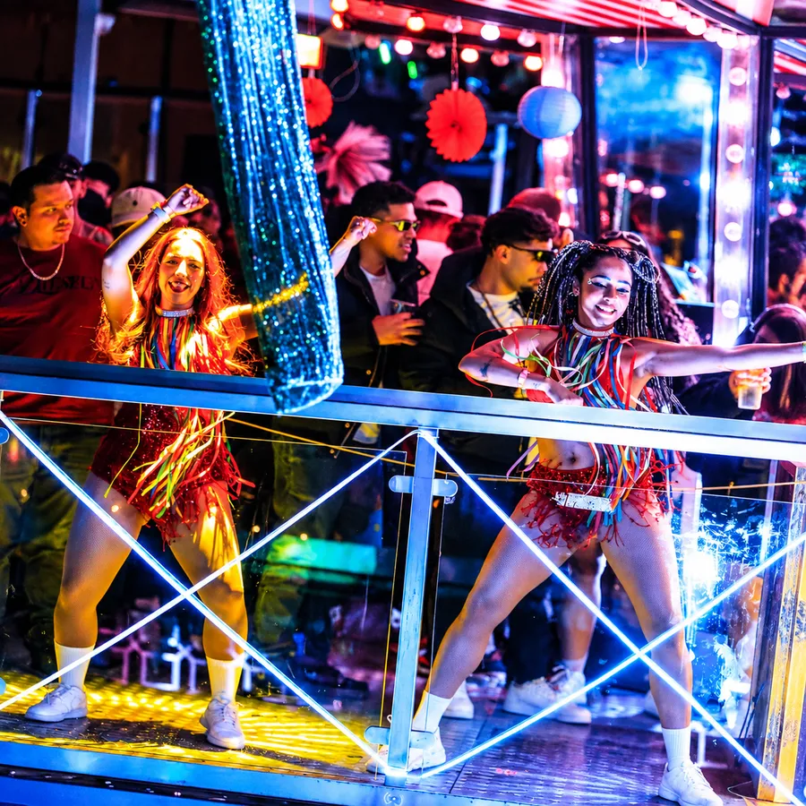 Two Brazil FitDance dancers in colourful ribbon costumes performing on a lit-up party bus stage